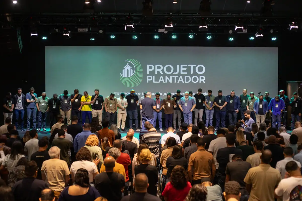 Plantadores reunidos no palco em oração durante a conferência, diante do telão do Projeto Plantador